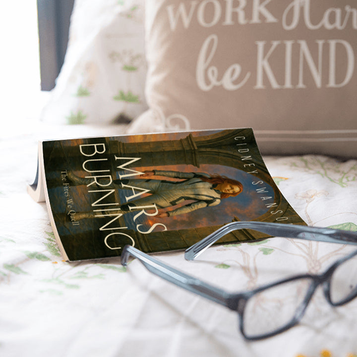 A copy of Mars Burning by Cidney Swanson — clean YA sci-fi for ages 10+, fourth in the Saving Mars series, next to glasses and a pillow.