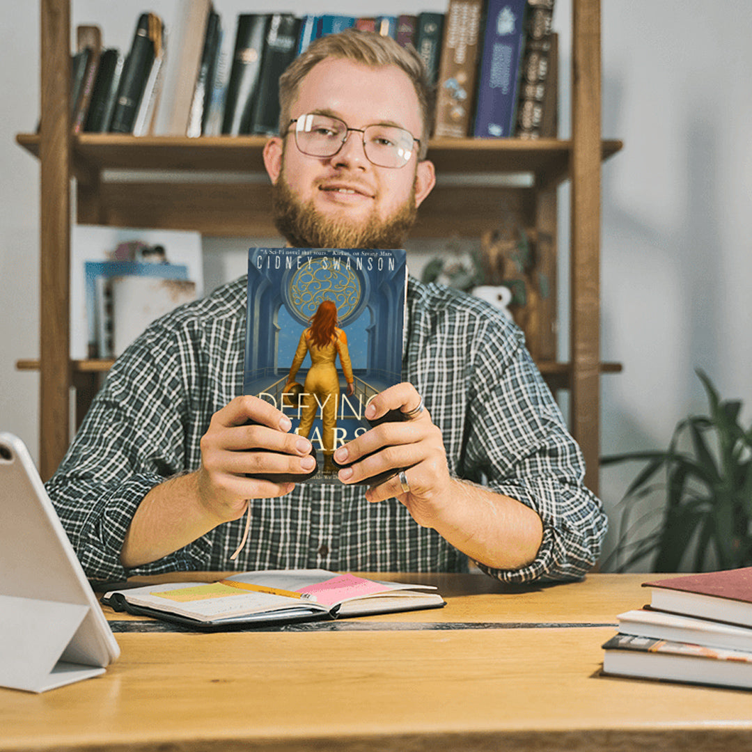 young man at desk holding Defying Mars by Cidney Swanson — clean YA sci-fi for ages 10+, second in the Saving Mars series.
