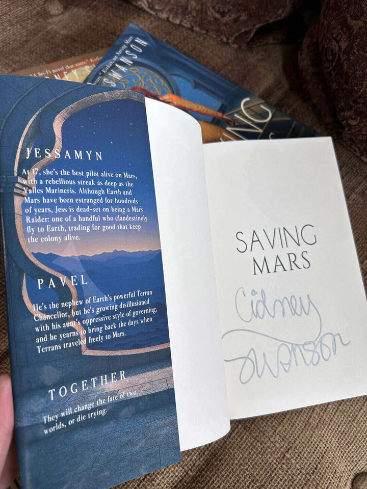 Blue book with text 'Saving Mars' and signed by author Cidney Swanson and held over a brown fabric background.