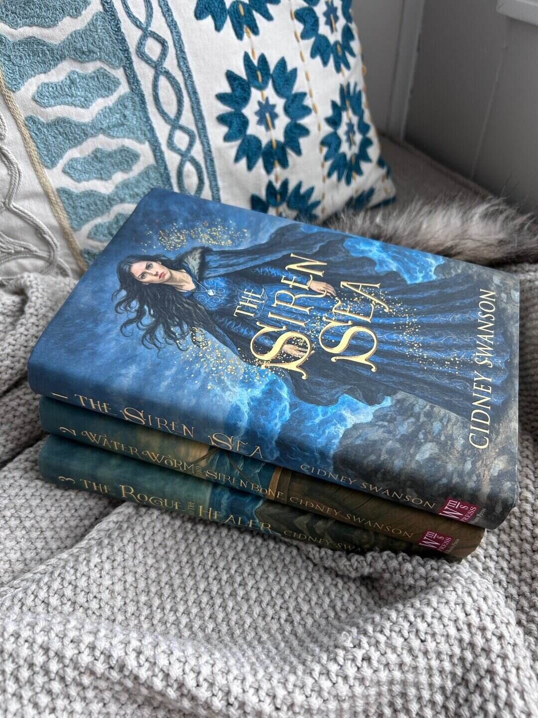 Stack of books by Cidney Swanson with The Siren Sea on top, laying on a blanket beside an embroidered blue and gold pillow